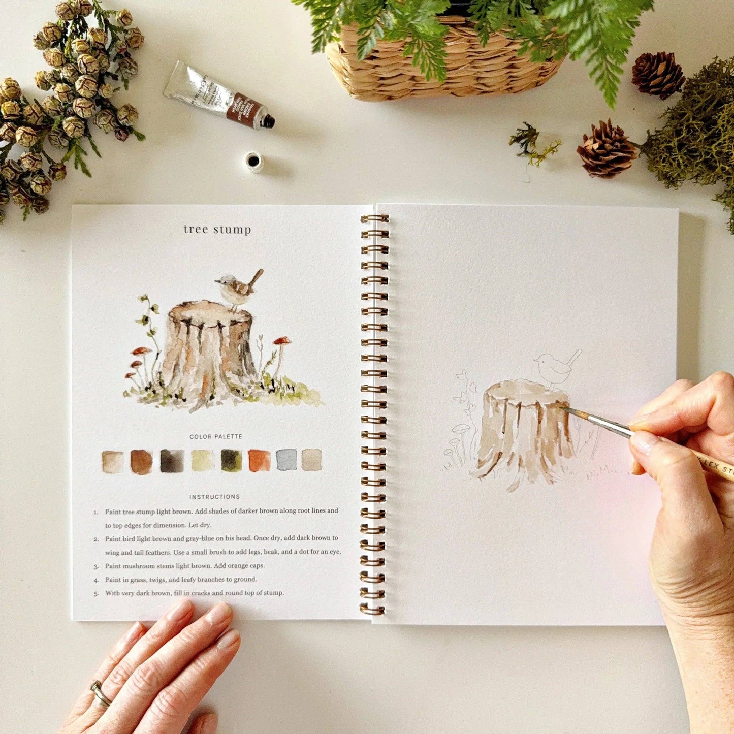 The Woodland Watercolor Workbook, available in standard size, is open to a page with a watercolor tree stump painting, color palette, and step-by-step instructions.