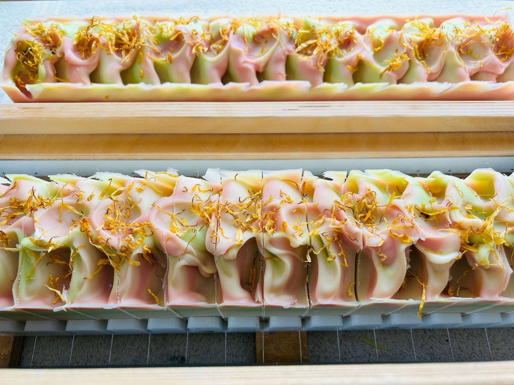 Handmade soap bars, known as Rows of Wildflowers, featuring a swirl pattern in pink, white and green colors. Each bar is topped with dried flower petals. The soaps are displayed on wooden racks. Available in one standard size.