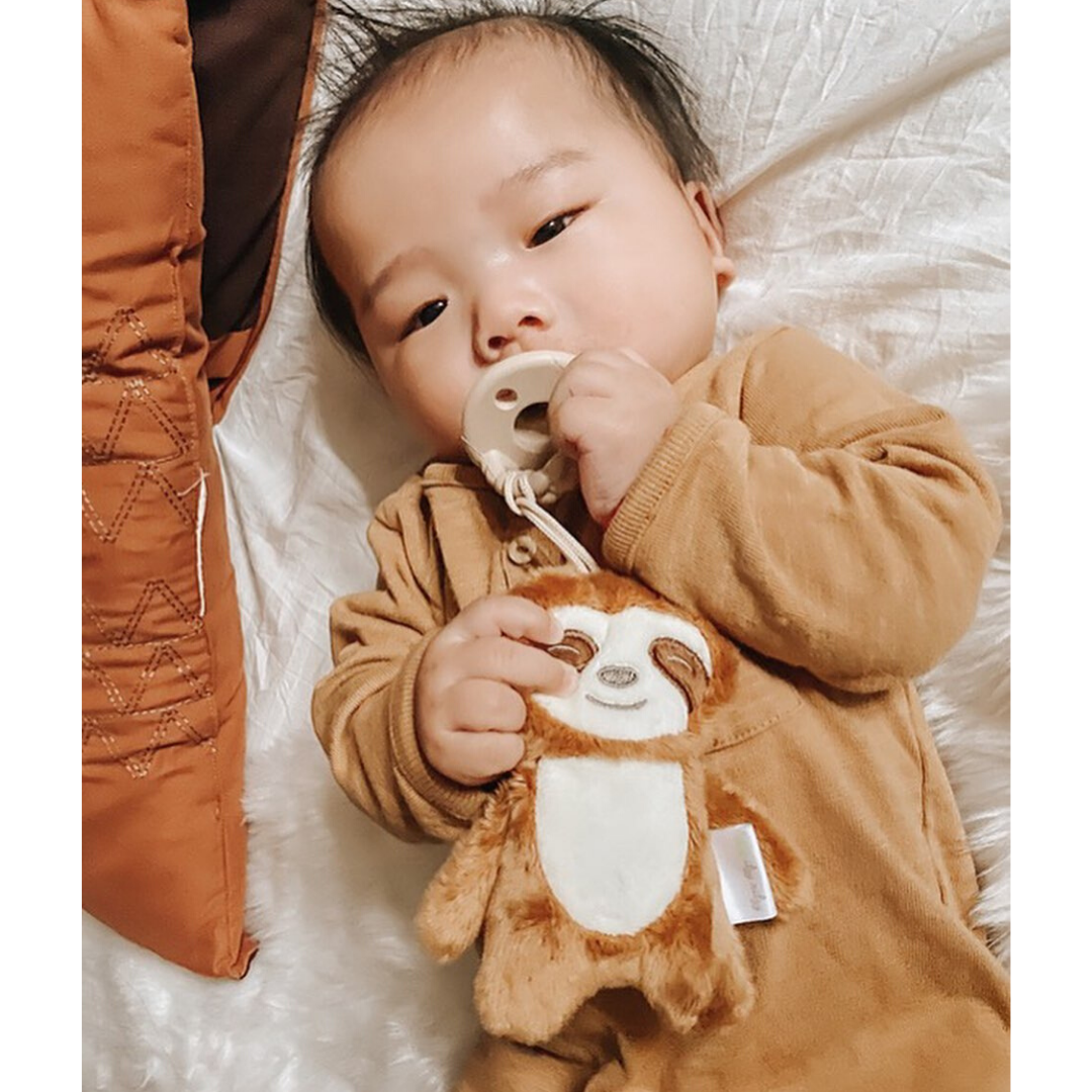 A baby wearing a cozy brown outfit lies on a white blanket, holding a Sweetie Pal™ Plush & Pacifier: Dino and a plush sloth toy. Pillows are on the left side.