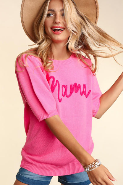 A joyful woman wearing a pink "Mama Embroidered Pop Letter Short Bubble Sleeve Sweater" with a broad-brimmed hat, swinging her blonde hair playfully. She's smiling broadly in a beige background.