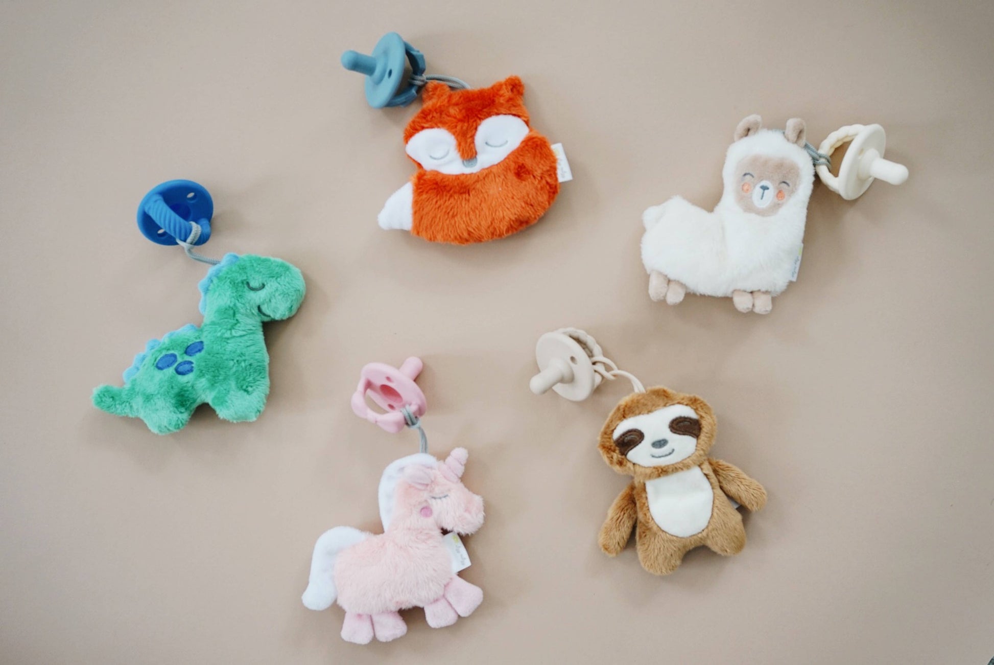 Various colorful plush toys, including a dinosaur, an orange fox, a llama, a Sweetie Pal™ Plush & Pacifier: Dino unicorn, a sloth, and a blue rattle, displayed on a light beige wall.