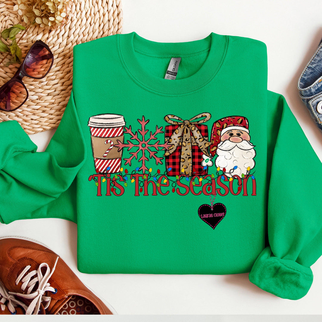 Light green “Tis The Season” sweatshirt featuring a coffee cup, snowflake, gift box, and Santa Claus graphic with “Tis The Season” text draped in string lights. Sizes available.