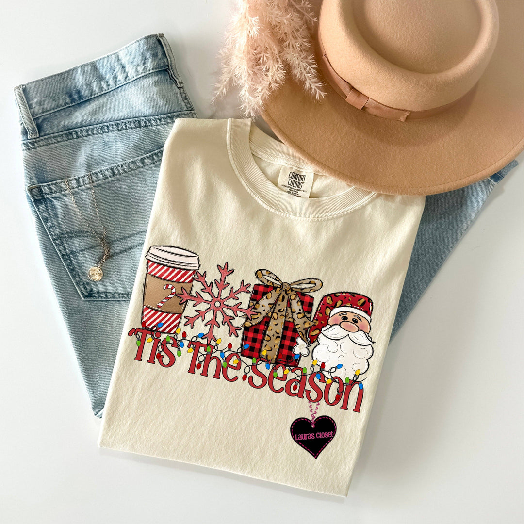 Cream “Tis The Season” graphic tee with a Christmas-themed coffee, snowflake, gift, and Santa design. Available in sizes S-XXL.