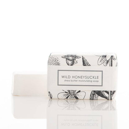 This product is a set of two bars of Wild Honeysuckle Soap - Bath Bar. The soap is made from shea butter, entirely vegan and features the aroma of wild honeysuckle. One bar comes unwrapped while the other is neatly packed in a package with floral designs. The soaps are placed on a reflective white surface for display purposes only. Available sizes not stated.