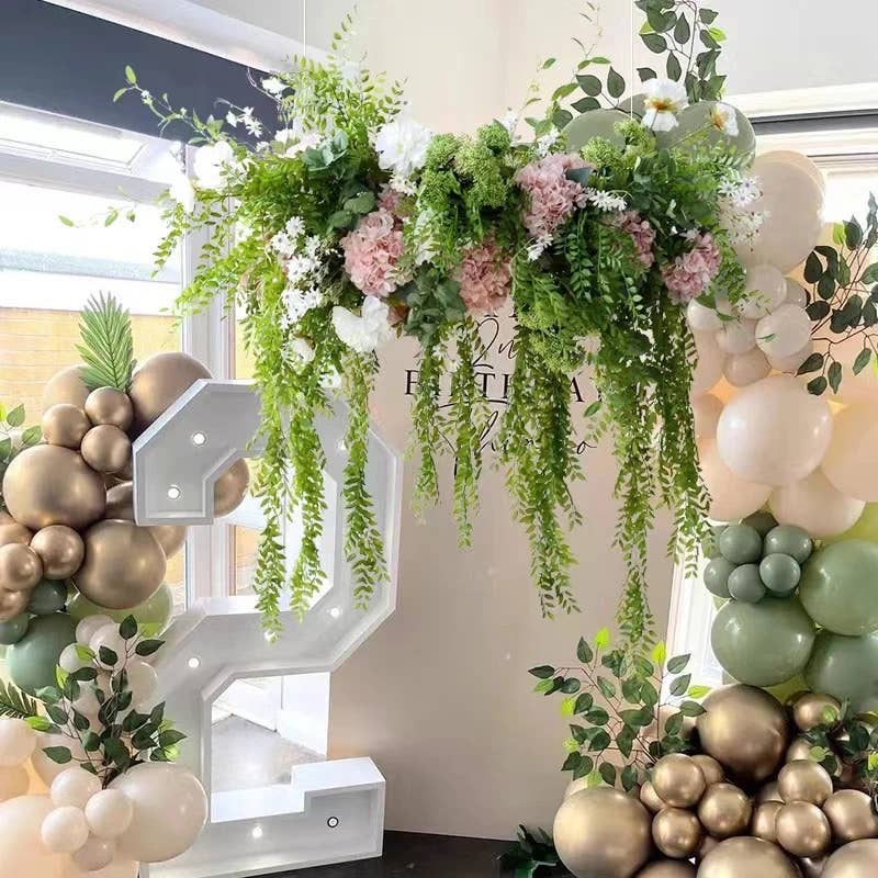 Decorative floral and balloon arch with lush greenery, white snowball flowers, pink blooms, and balloons in cream, gold, and green. Includes artificial botanicals. Features illuminated white "C" surrounded by more balloons. Showcases 40" Long-5 Blooms-Premium Faux Snowball hydrangea spray: Green.