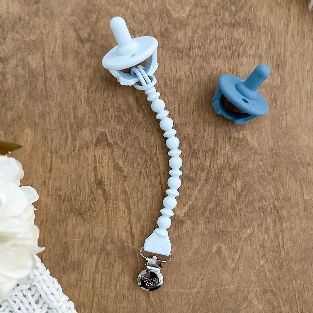 Two pacifiers on a wooden surface, one attached to a white Sweetie Strap™ Silicone One-Piece Pacifier Clips: Hero Blue Beaded with a metal clasp, and another blue pacifier nearby. A soft focus flower is partially