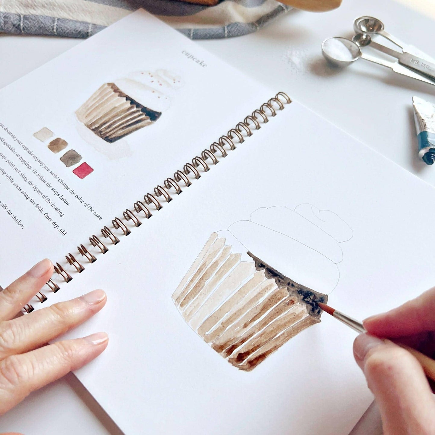 A hand uses a paintbrush to color a cupcake illustration in the Baking watercolor workbook. Available sizes: 8x10 and 11x14 inches.