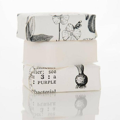 Gardenia Blossoms Soap - Bath Bar is a set of three stacked soap bars. Each bar is individually wrapped in paper featuring floral and botanical prints, with a prominent gardenia blossom design. The soap bars come attractively reflected on a white surface.