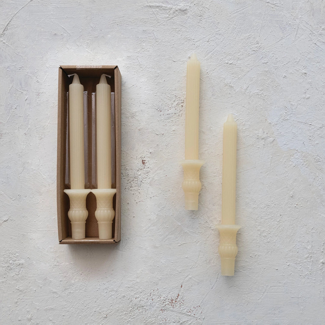 Brushed concrete background with set of 2 candles in box and set of 2 candles by themselves. Ivory candlesticks with urn bases