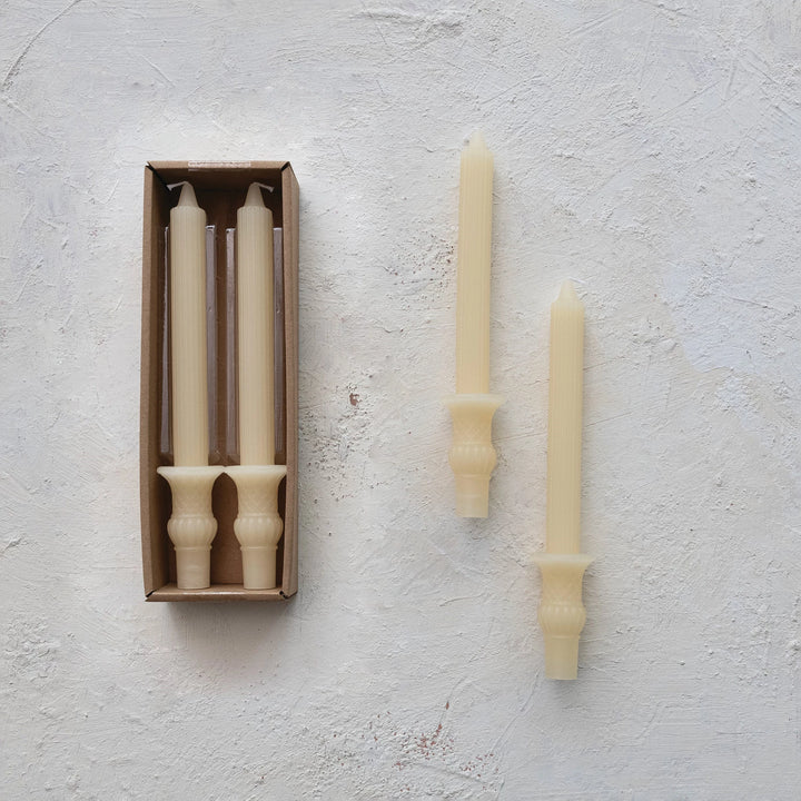 Brushed concrete background with set of 2 candles in box and set of 2 candles by themselves. Ivory candlesticks with urn bases