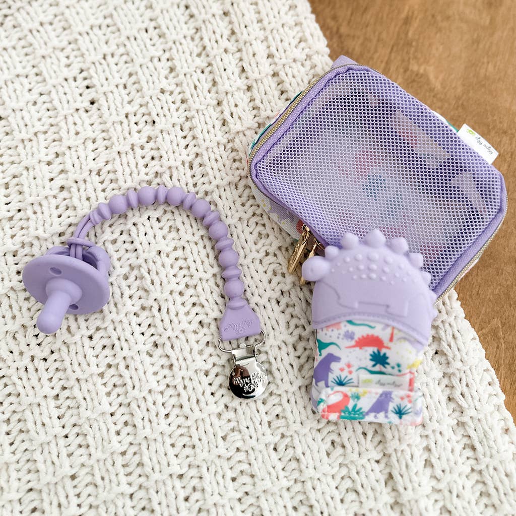 A purple baby teething toy and case on a white knit blanket next to a purple mesh zipper pouch, with a small sanitizer holder featuring a colorful floral pattern and a Sweetie Strap™ Silicone One-Piece Pacifier Clip: Hero Blue Beaded