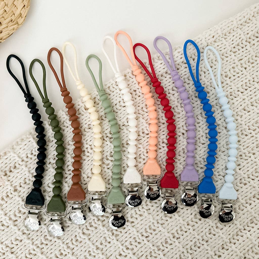 An array of Hero Blue Beaded Sweetie Strap™ Silicone One-Piece Pacifier Clips placed in a neat row on a cream crochet blanket. Each Sweetie Strap™ features a different color combination and is attached to a silver clasp.