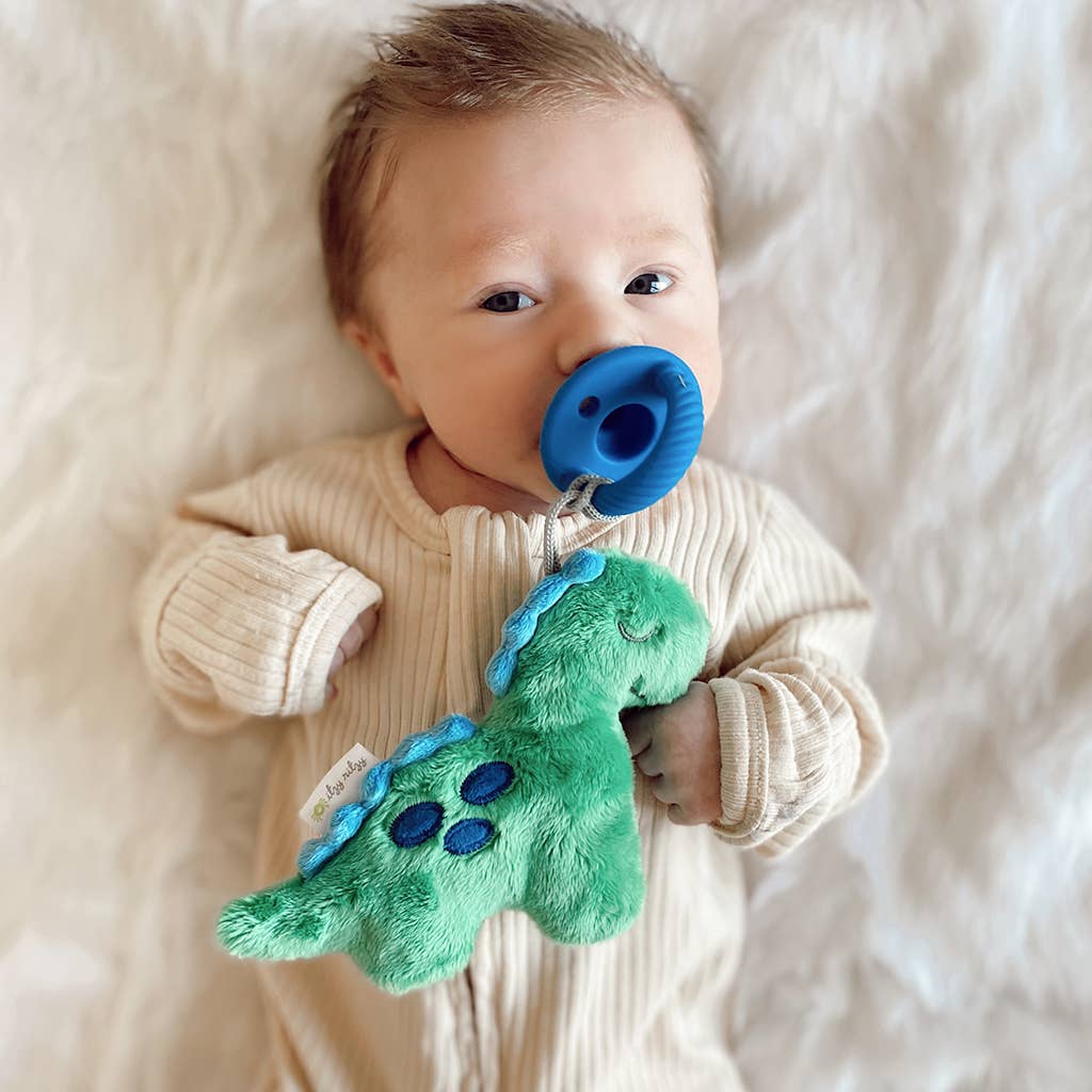 A newborn baby with a Sweetie Pal™ Plush & Pacifier: Dino in their mouth lies on a soft, white blanket. The baby is holding a green and blue dinosaur plush toy.