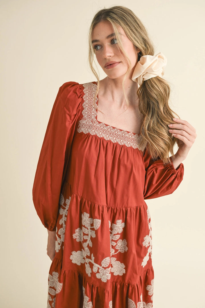 Rust midi dress with square neckline, cream lace trim, and cream floral embroidery. Available in all sizes.