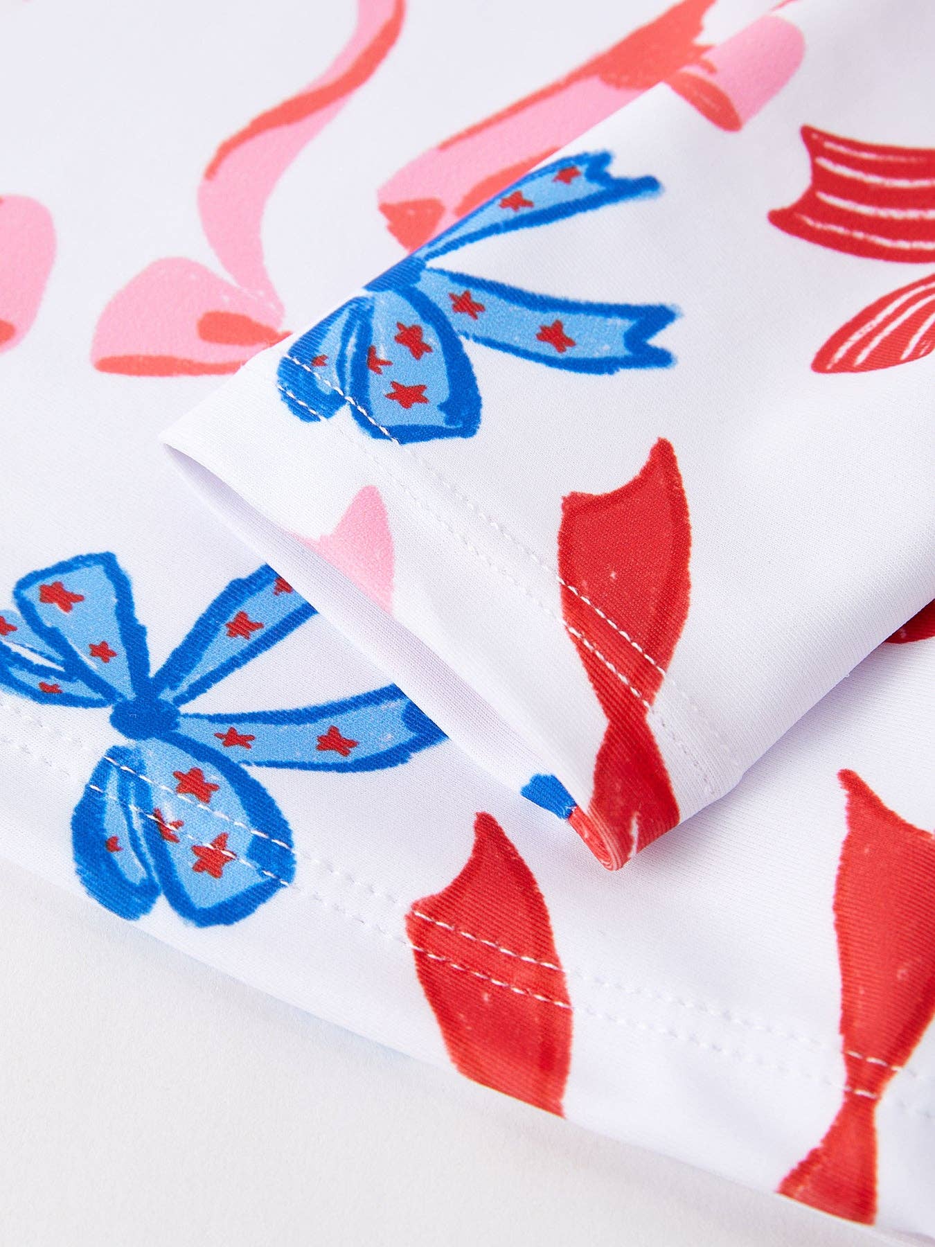 White swimsuit with blue bows, red stars, pink shapes, folded sleeve. Available in sizes XS-XL.