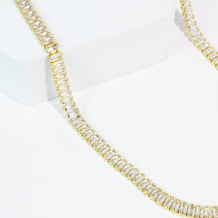 The Royale Necklace features a gold chain with rectangular clear gemstones and crystal accents. Available in 16" and 18" sizes.
