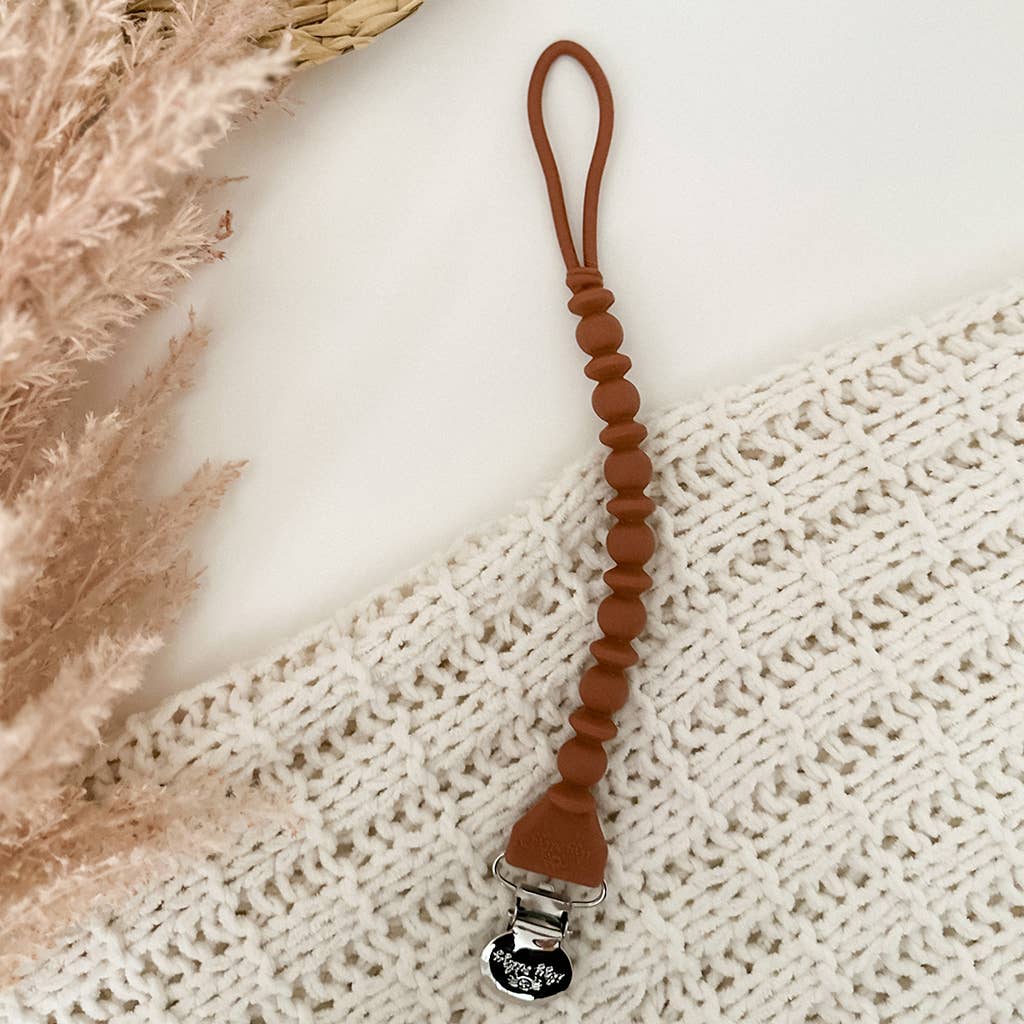 A Sweetie Strap™ Silicone One-Piece Pacifier Clips: Mint Braid laid on a textured white knit blanket with dried pampas grass in the background, conveying a soft and serene nursery ambiance.