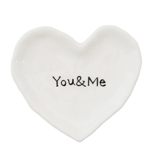 The You & Me Ceramic Heart Dish is a white, heart-shaped ceramic dish with "You & Me" inscribed at the center in black letters. Ideal for holding spare change or rings. Available in one size.