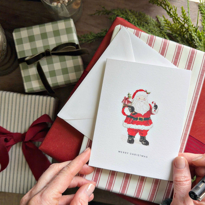 Watercolor Santa greeting card with sack of toys, candy cane, and “MERRY CHRISTMAS” text. Available in multiple sizes.