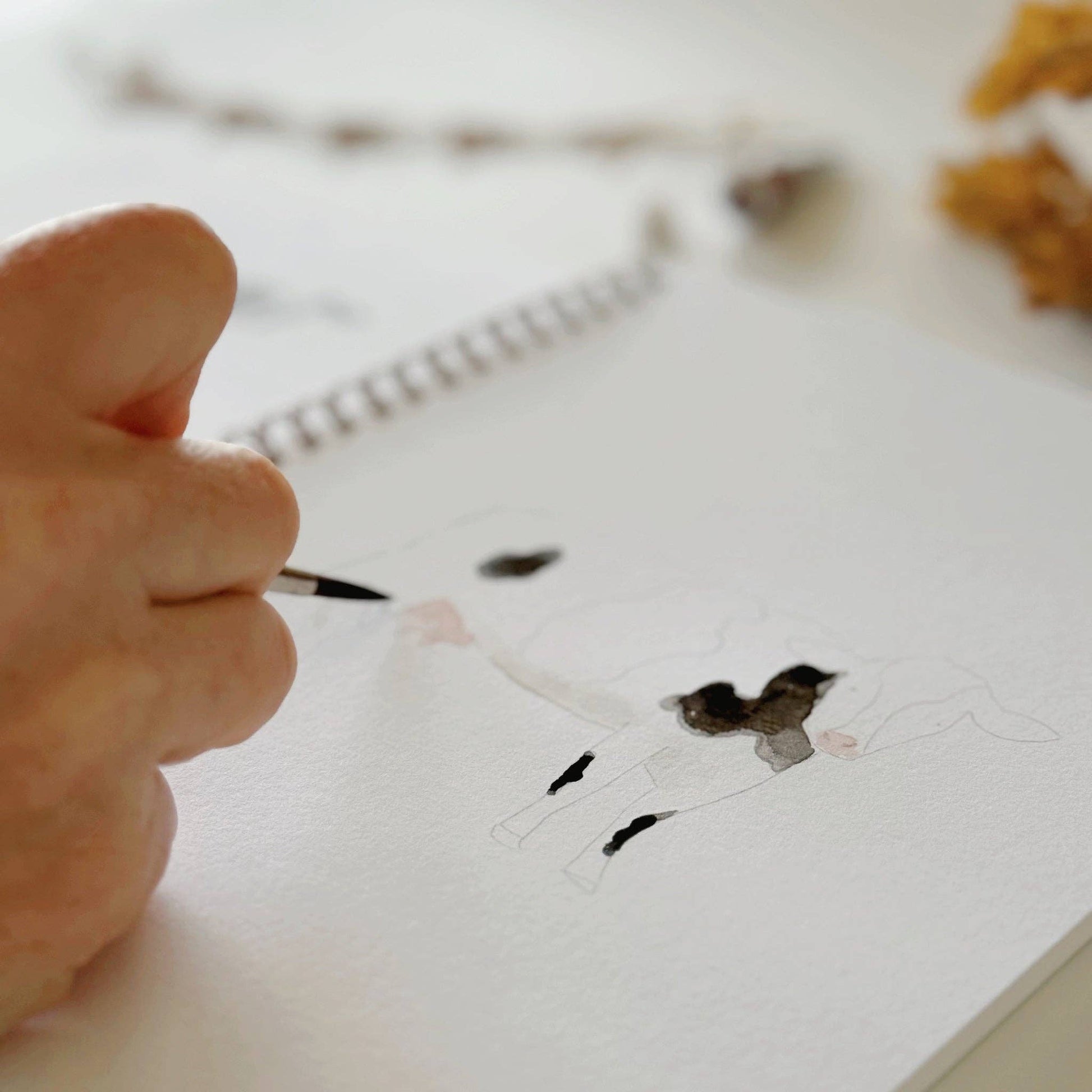 A hand paints a black and white calf in the Farm Watercolor Workbook sketchbook. Available sizes: 8x10 and 9x12 inches.