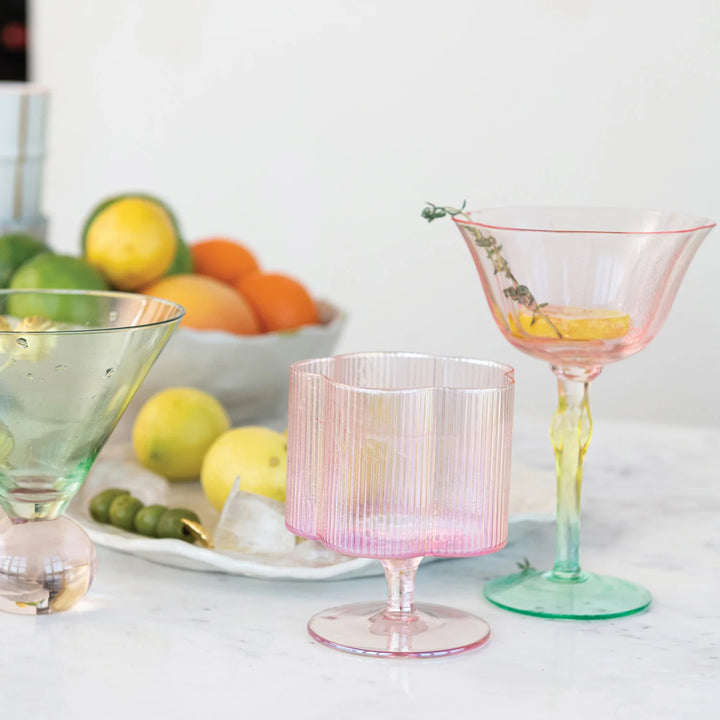 Colorful glassware featuring a pink goblet, a hand-blown pleated flower-shaped wine glass with an iridescent finish, and ribbed pink glass. Sizes available: standard drinkware dimensions.