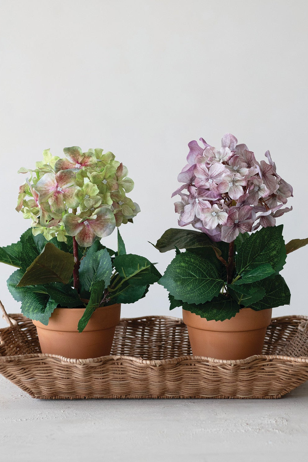 Two faux hydrangea arrangements, one pale green and one light purple, feature realistic petals in terracotta-style plastic pots. Available in small and medium sizes.