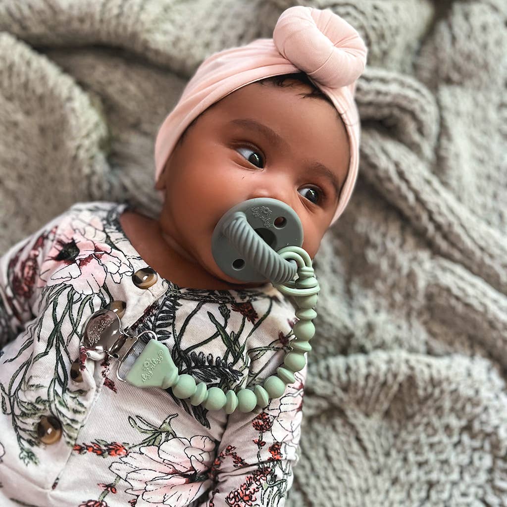 A baby with a Sweetie Strap™ Silicone One-Piece Pacifier Clip lies on a textured blanket, wearing a floral outfit and a pink headband styled like a bow.