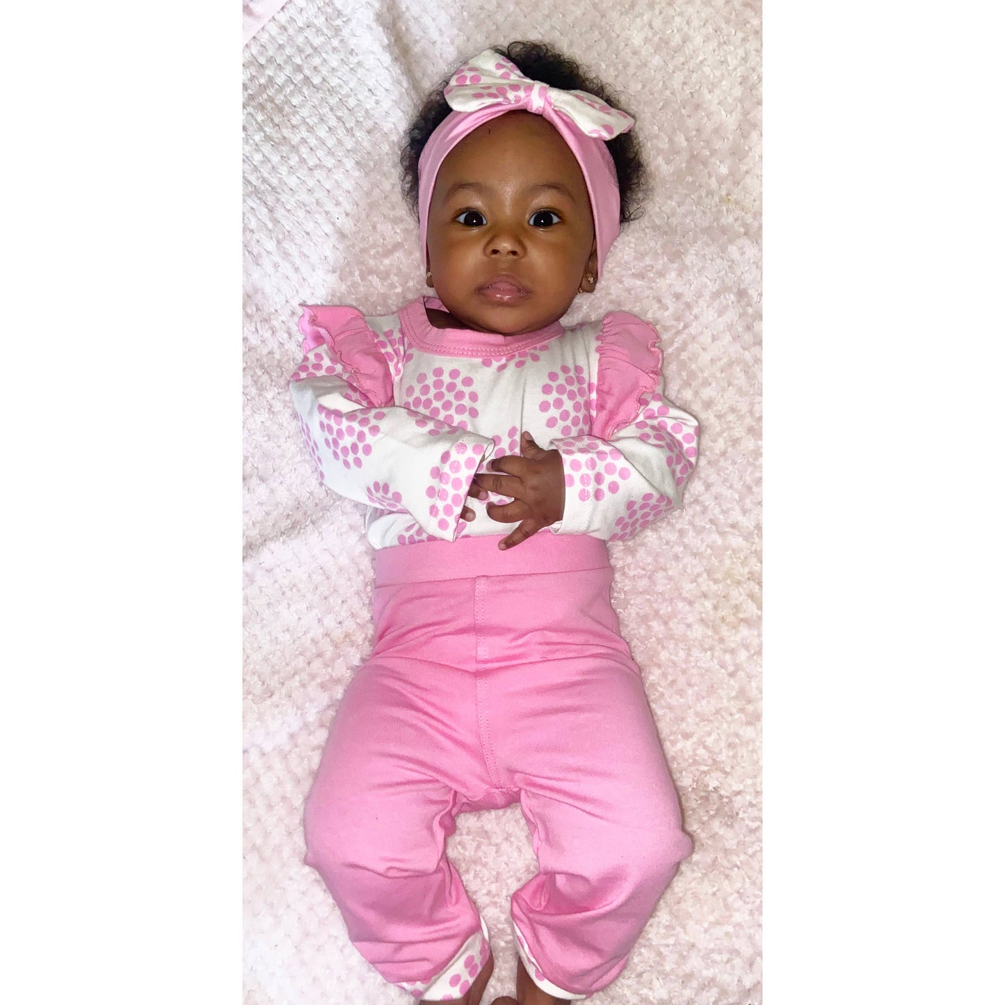 A baby girl dressed in an Layette Girls Baby clothes Shower Gift Pink Polka Dot Easter: 3-6 Mo outfit with polka dot patterns and a matching headband lies happily on a soft white surface, looking up at the camera.