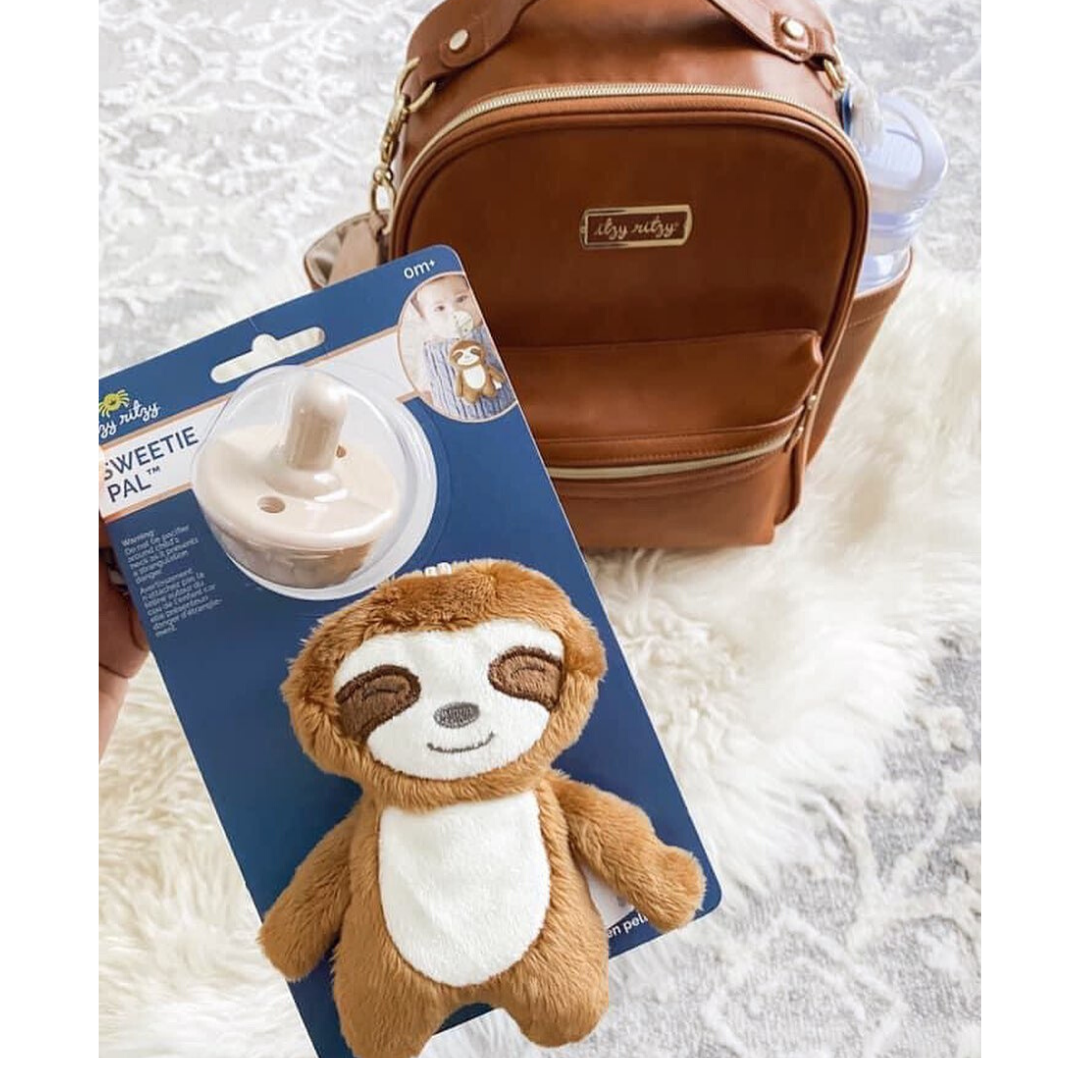 A hand holds a Sweetie Pal™ Plush & Pacifier: Dino packaged with a plush sloth, above a brown diaper bag on a white furry surface.