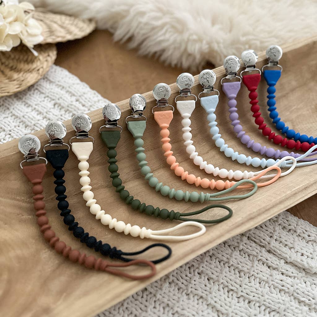 A wooden tray displaying a collection of colorful Sweetie Strap™ Silicone One-Piece Pacifier Clips: Hero Blue Beaded with metal clasps, arranged neatly on a soft, textured background.