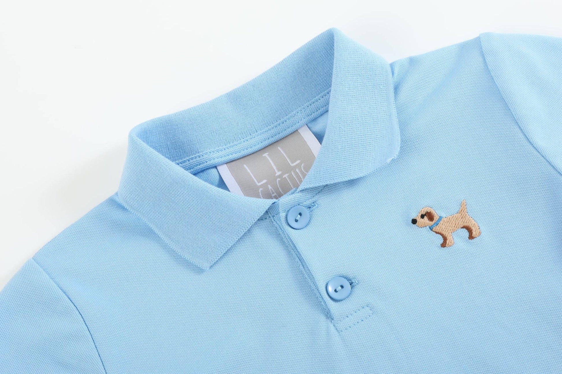 Light blue polo shirt with collar, two buttons, dog with sunglasses patch; white inner tag. Sizes: XS, S, M, L, XL.