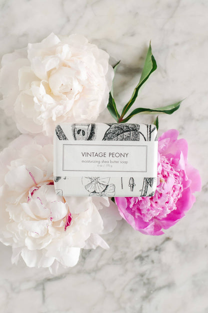 This Vintage Peony Soap is a bath bar presented in beautiful floral paper wrapping. It carries the delicate scent of lush white and pink peony flowers. The soap has been crafted to capture the aesthetic of a vintage era, resting against an elegantly polished marble surface. The product is available in standard soap bar size.