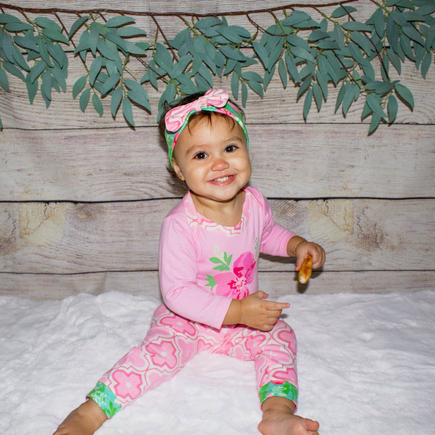 Older baby modeling set sitting in front of a wood background with greenery. She is smiling and eating some pizza crust.