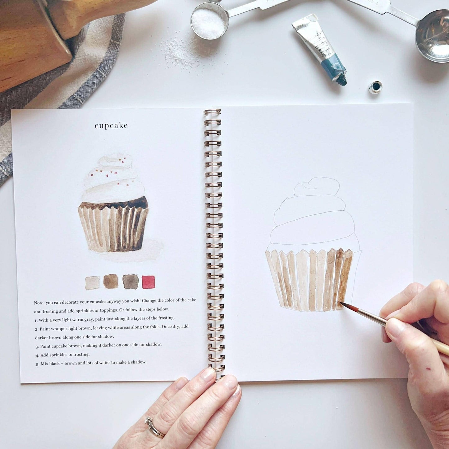The Baking watercolor workbook is a spiral-bound art book featuring baking illustrations, color swatches, and step-by-step instructions; available in multiple sizes.