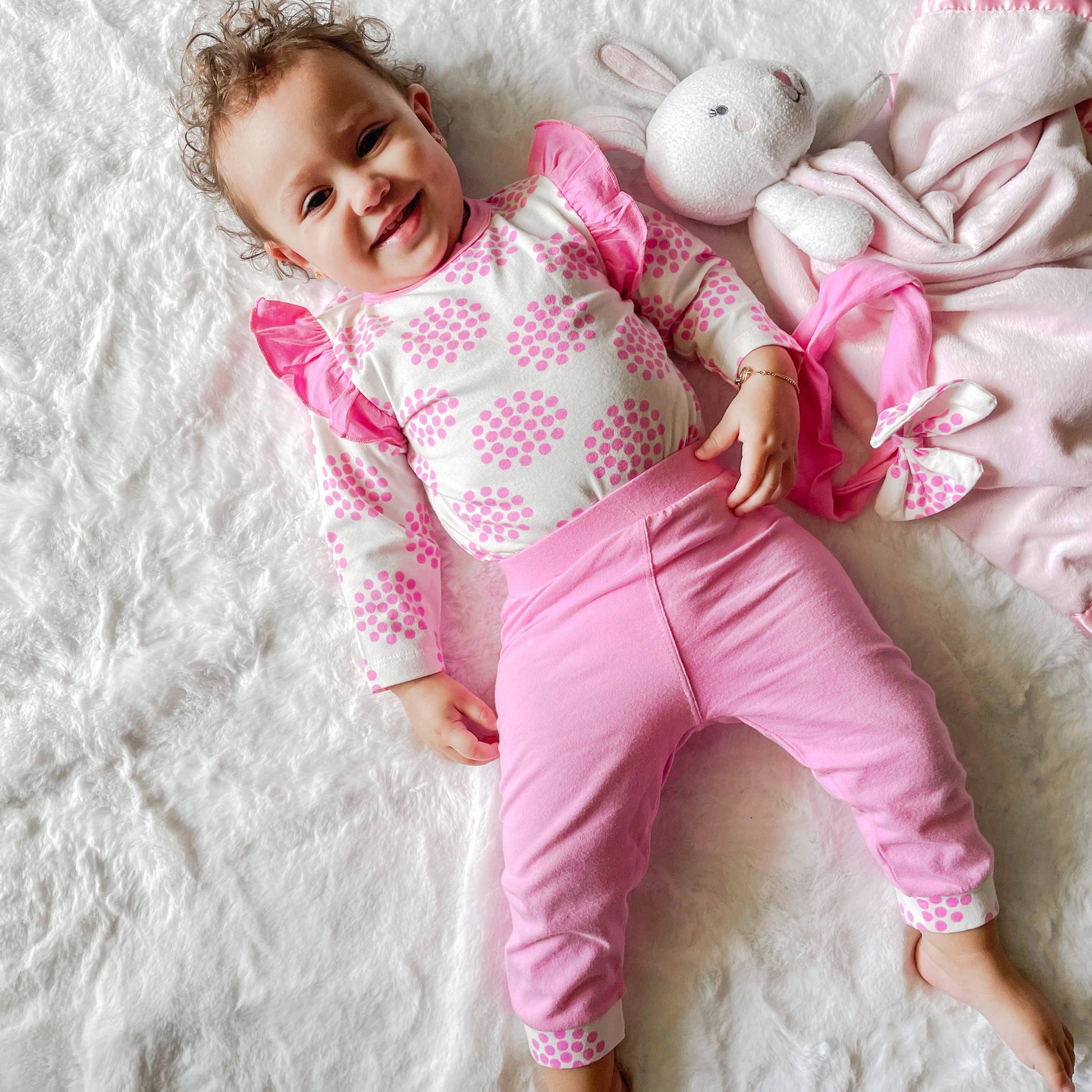 A smiling baby with curly hair lying on a white fuzzy blanket, wearing the Layette Girls Baby clothes Shower Gift Pink Polka Dot Easter: 3-6 Mo outfit from AnnLoren design, next to a plush rabbit toy.