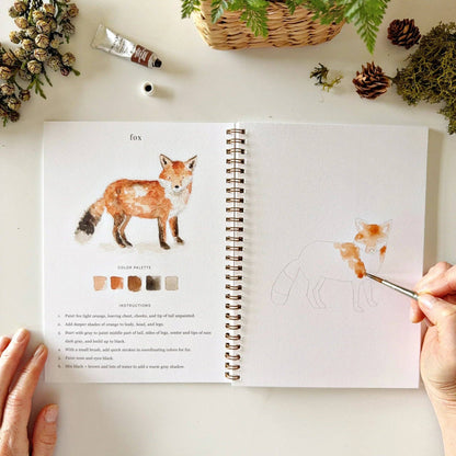 The Woodland watercolor workbook is open to a fox painting tutorial. Available in multiple sizes.
