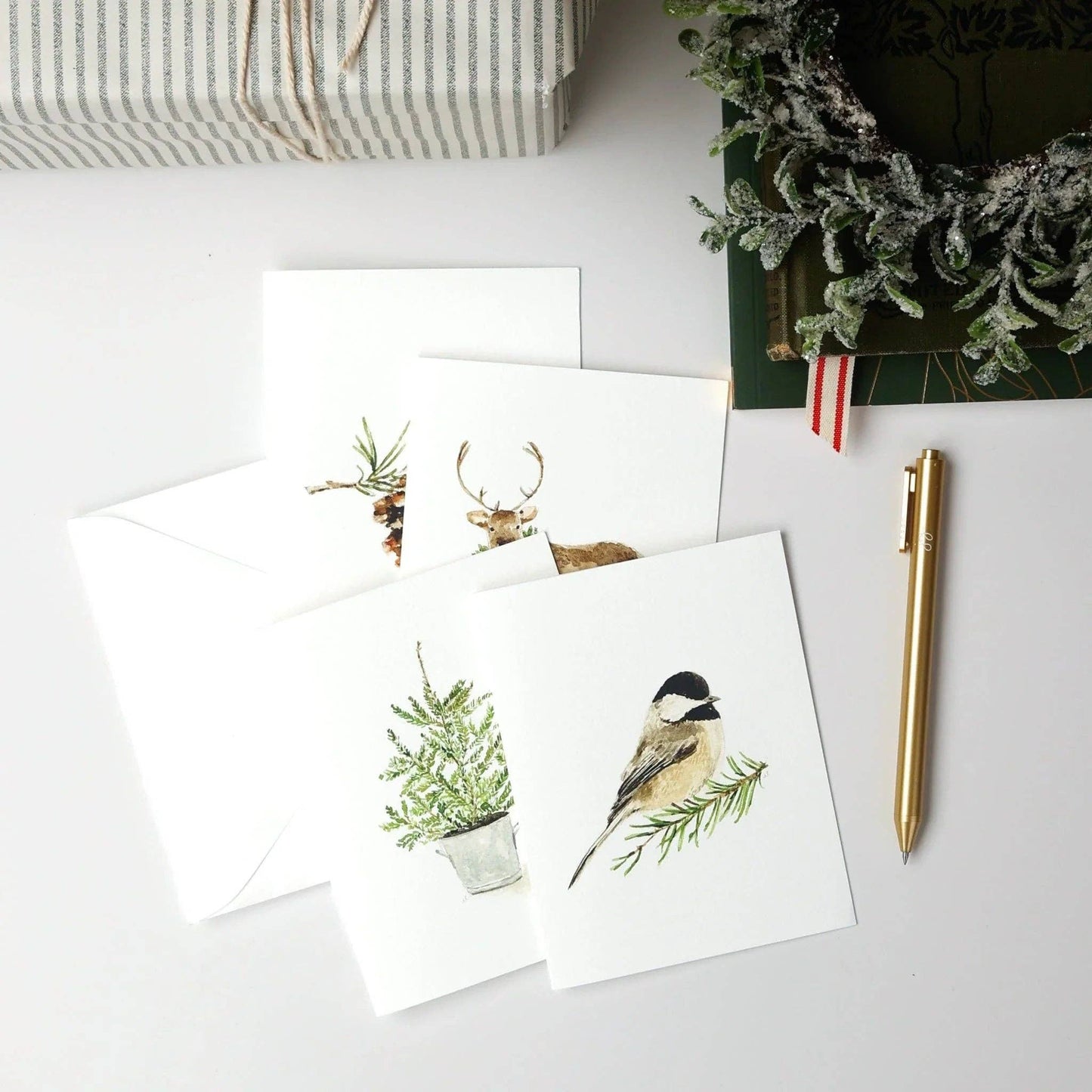 The Evergreen Christmas assorted notecard set includes four white watercolor cards with delicate animal and plant designs. Available in standard notecard size.