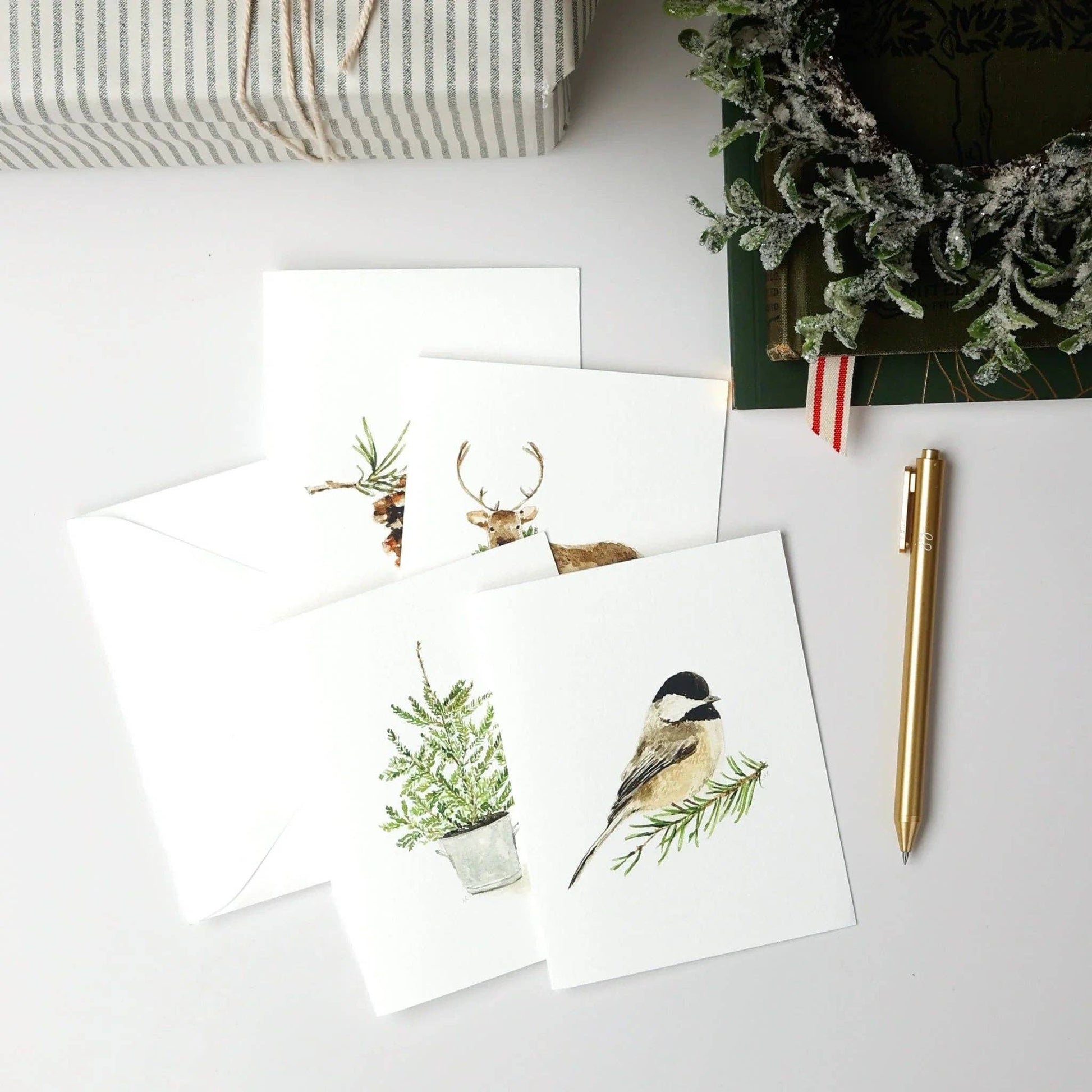 The Evergreen Christmas assorted notecard set includes four white watercolor cards with delicate animal and plant designs. Available in standard notecard size.