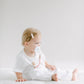 A toddler with blonde hair, wearing a simple white outfit, sits on a white surface holding a BPA free Pacifier Clip - Pink and Blue, looking thoughtfully to the side in a brightly lit room.