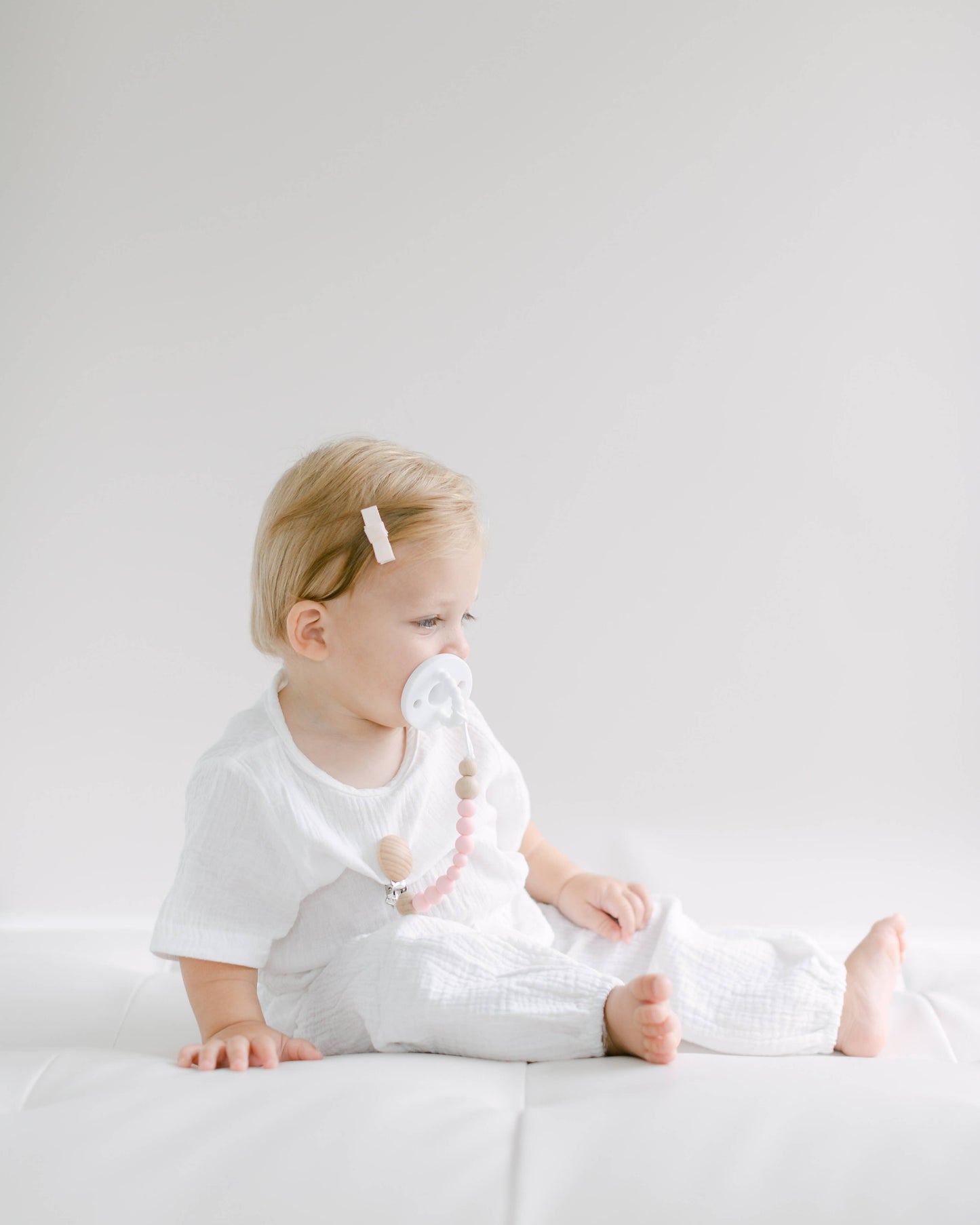 A toddler with blonde hair, wearing a simple white outfit, sits on a white surface holding a BPA free Pacifier Clip - Pink and Blue, looking thoughtfully to the side in a brightly lit room.