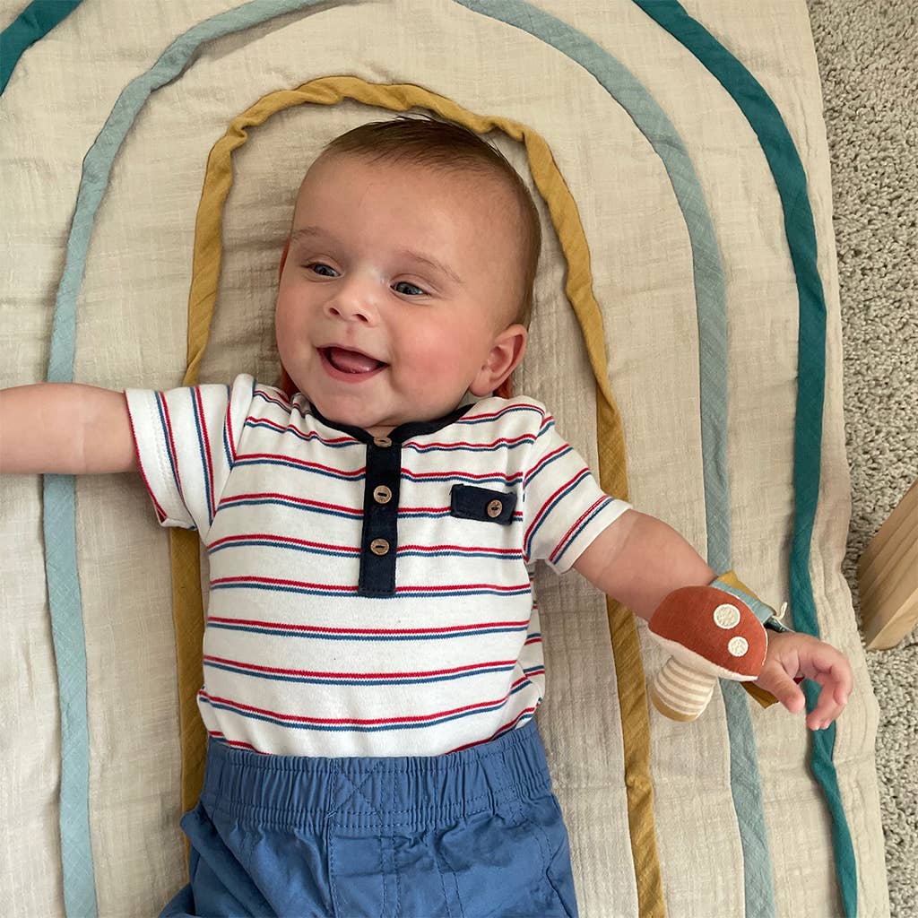 A joyful baby in a striped shirt and blue shorts lying on a colorful circular play mat, smiling and reaching out playfully with an Itzy Bitzy Wrist Rattle: Cottage.