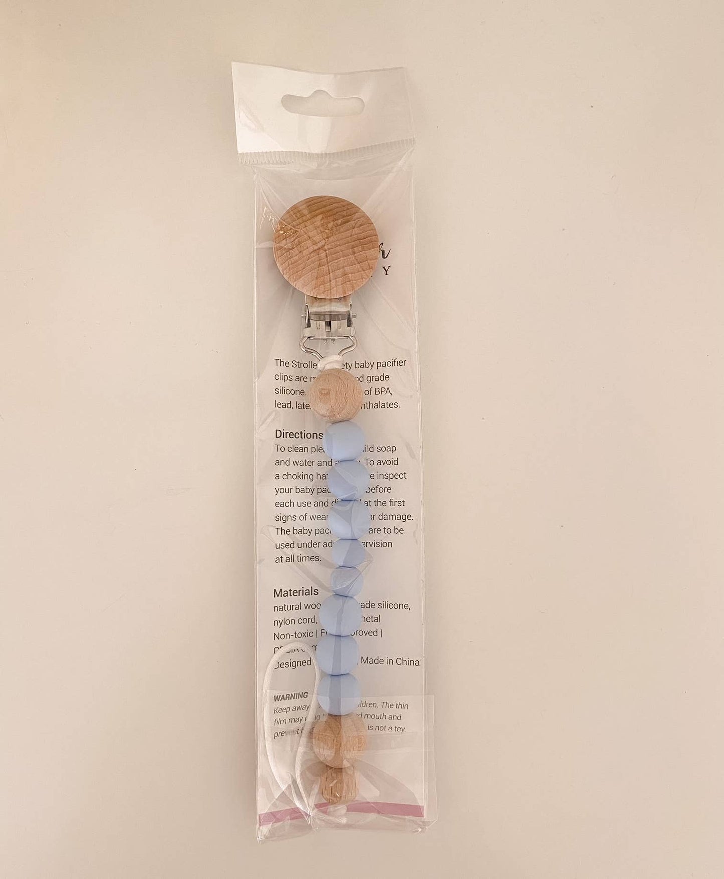 A Pacifier Clip - Pink and Blue with a BPA-free, wooden circular clip and food-grade silicone beads, packaged in a clear, plastic hanging package with text describing the product's features and usage instructions.