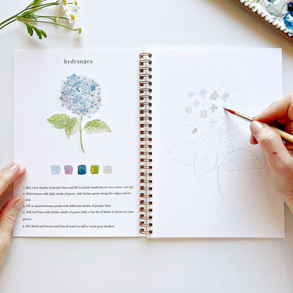 A hand paints blue flowers in the Flowers Watercolor Workbook, which has finished samples and instructions. Sizes: Standard.