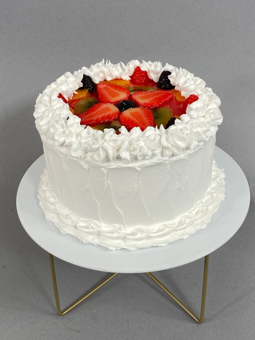 Small fake vanilla cake topped with fruit, white frosting, piped cream, strawberries, kiwi, berries. Available in small size.
