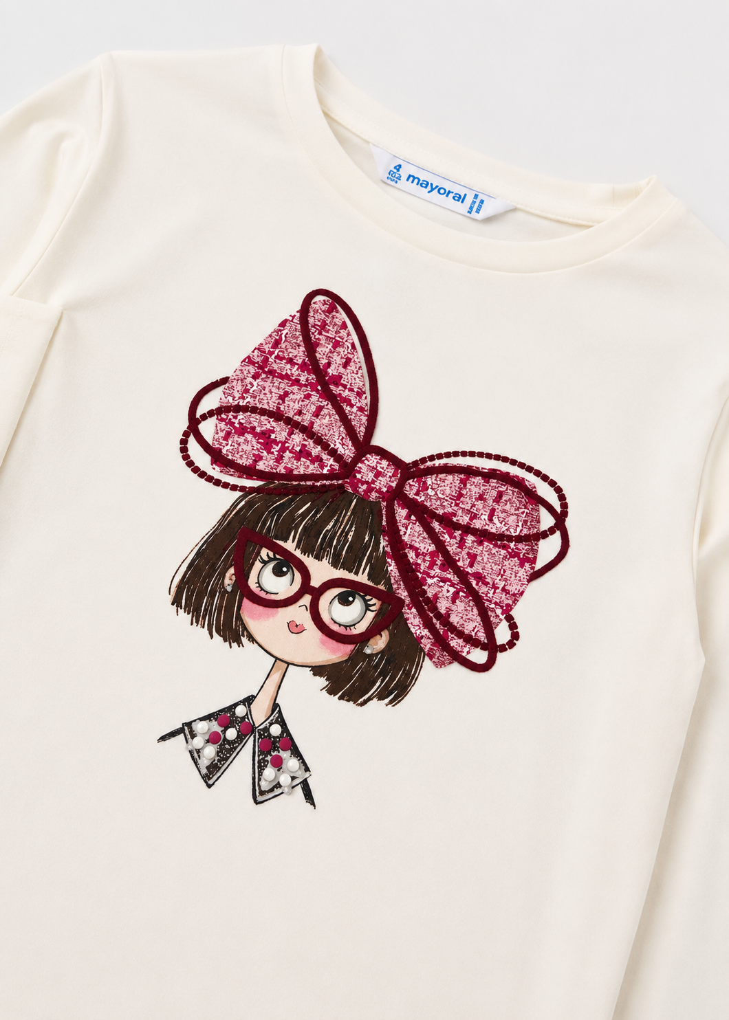 Cream-colored long sleeve t-shirt for girls with an illustration of a girl wearing red glasses and a pink bow, decorative gem collar, and “mayoral” label. Available in sizes 2-8 years.