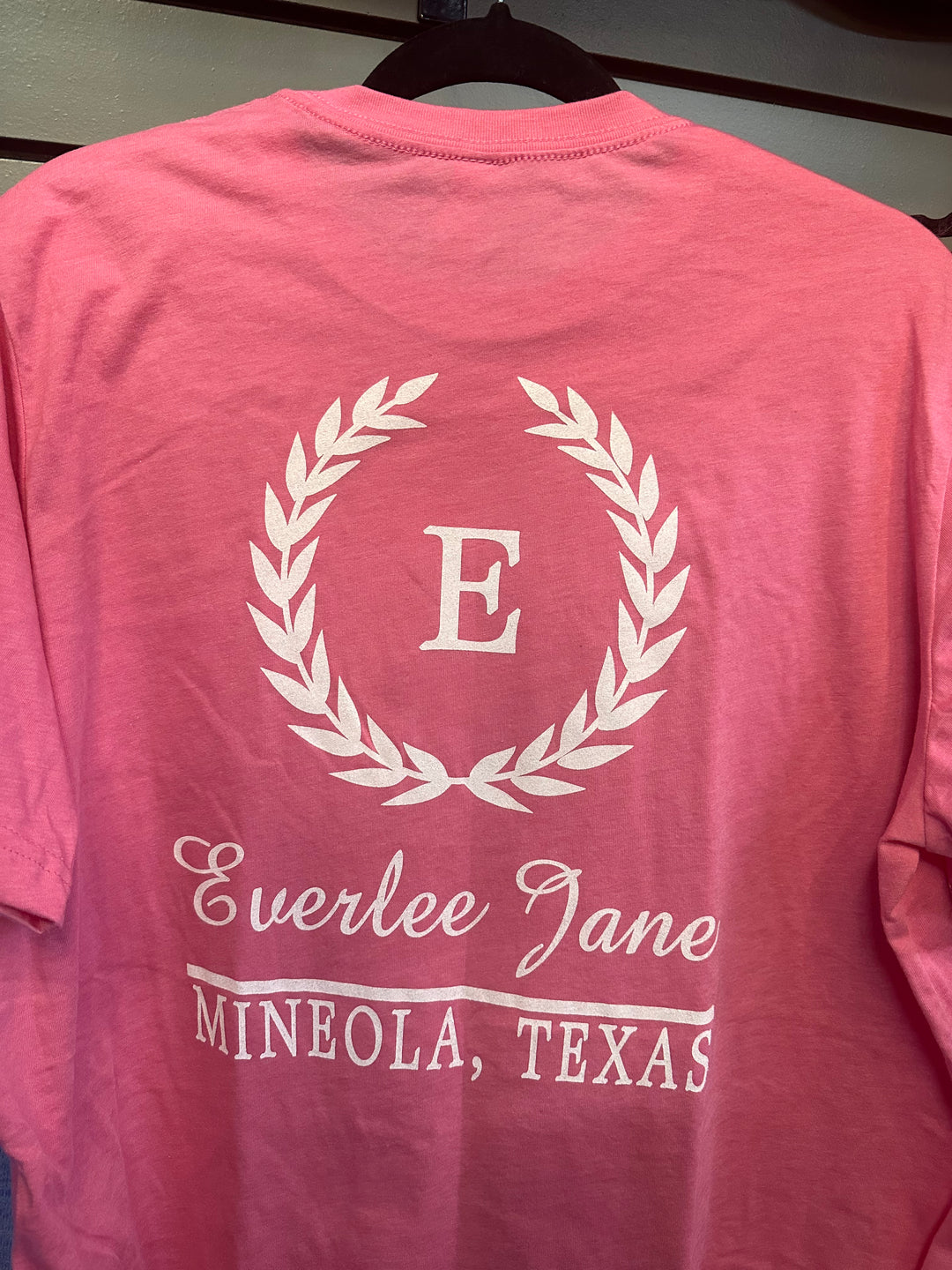 A pink Everlee Jane Tshirt hanging on a hanger with a white printed design featuring a wreath and the initial "e" in the center. Below the wreath, the text reads "Ever".