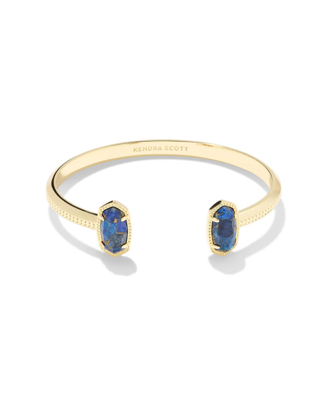 The Elton Gold Cuff Bracelet by Kendra Scott features Bronze Veined Blue Lapis stones on each end, a textured edge, and “Kendra Scott” engraved inside. Adjustable open cuff design. Available in one size.