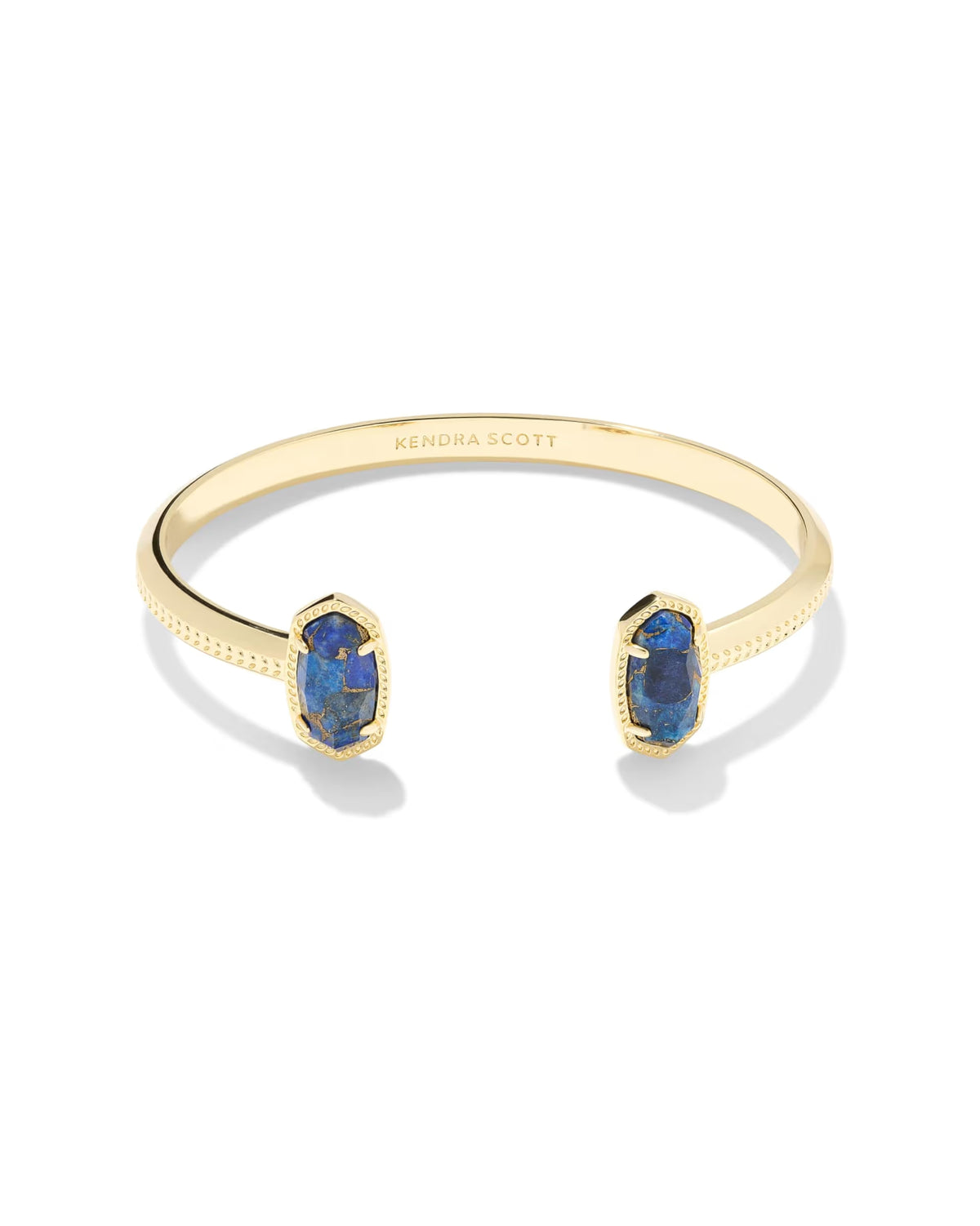 The Elton Gold Cuff Bracelet by Kendra Scott features Bronze Veined Blue Lapis stones on each end, a textured edge, and “Kendra Scott” engraved inside. Adjustable open cuff design. Available in one size.