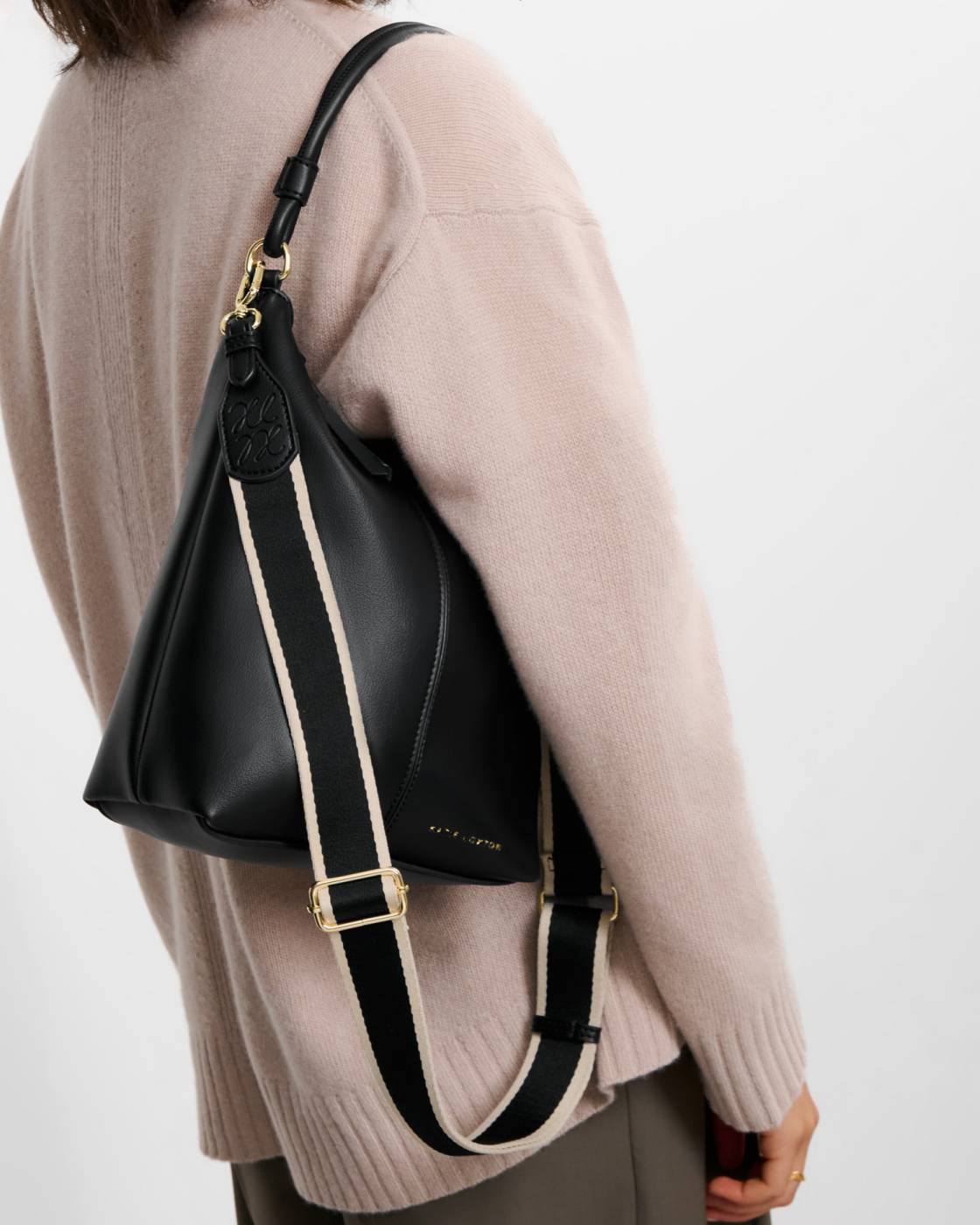 Hallie Satchel Bag in black vegan leather with gold hardware, embossed logo near the top, and a wide black and white shoulder strap; available in small, medium, and large sizes.