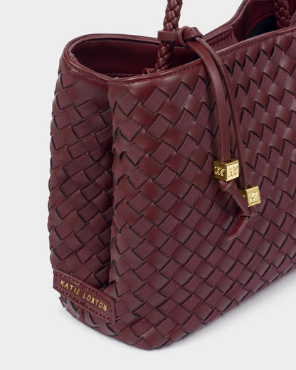 Nova Hand Woven Top Handle Bag in Cherry, made of maroon woven vegan leather with gold cube charms on the drawstring and a gold “Katie Loxton” label. Includes top handle, crossbody strap. Available in one size.